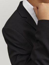 Svart kavaj med synlig sömnad och textur; skarp vit skjortkrage. Detaljerna av tyg och skrädderi är framträdande.