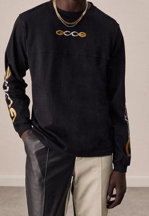 Mann trägt ein schwarzes Langarmshirt mit gold- und weißem Kettendesign auf Brust und Ärmeln, kombiniert mit zweifarbigen schwarzen und beigen Hosen.