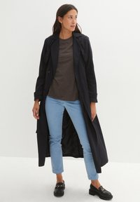 Cappotto trench blu navy con dettagli di bottoni, indossato sopra una maglia marrone, abbinato a jeans azzurri chiari e mocassini neri con un accento di catena dorata.