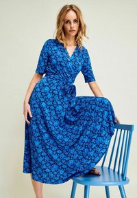 Sininen kukkakuosiset wrap-mekko, jossa on lyhyet hihat ja vyötäröllä solmittava nauha, ja joka sisältää valuva hame, istuen sinisellä tuolilla.