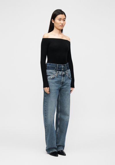 Μαύρο τοπ με μακριά μανίκια και ανοιχτούς ώμους, συνδυασμένο με ψηλόμεσα, φαρδιά τζιν παντελόνια ανοιχτού μπλε χρώματος. Μαύρα παπούτσια με μυτερή μύτη. Απλός, μινιμαλιστικός σχεδιασμός.