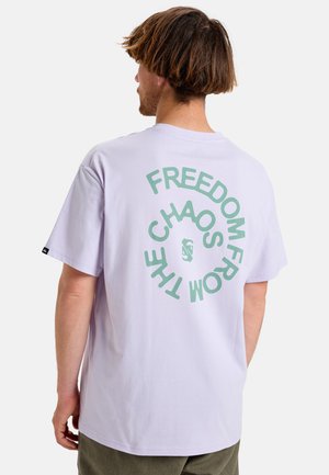 Man gezien van achteren, die een lichtpaarse T-shirt draagt met groene tekst waarop "Freedom from the chaos" staat in een cirkelvormig patroon.