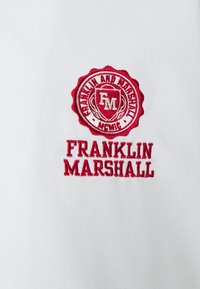 Červeně vyšitý logo a text Franklin and Marshall na bílé látce, s emblemem ve středu obsahujícím iniciály "FM".