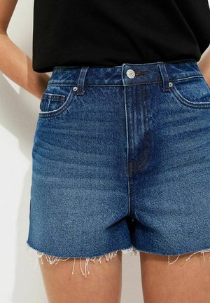 Person wearing high-waisted blue denim shorts with frayed hems and a black tucked-in top, standing against a light background.