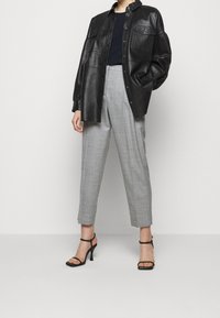 Černá kožená oversized košile-bunda se dvěma kapsami na hrudi, doplněná světle šedými kalhotami na míru a černými sandály na podpatku.