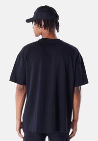 T-shirt en coton noir surdimensionné à manches courtes, avec un col ras du cou côtelé et une texture lisse ; porté avec une casquette noire assortie.