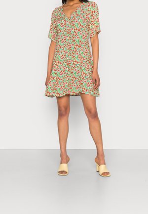 Frau trägt ein kurzes, geblümtes Kleid mit kurzen Ärmeln und V-Ausschnitt, kombiniert mit beigen, eleganten Slip-On-Sandalen mit Absatz.