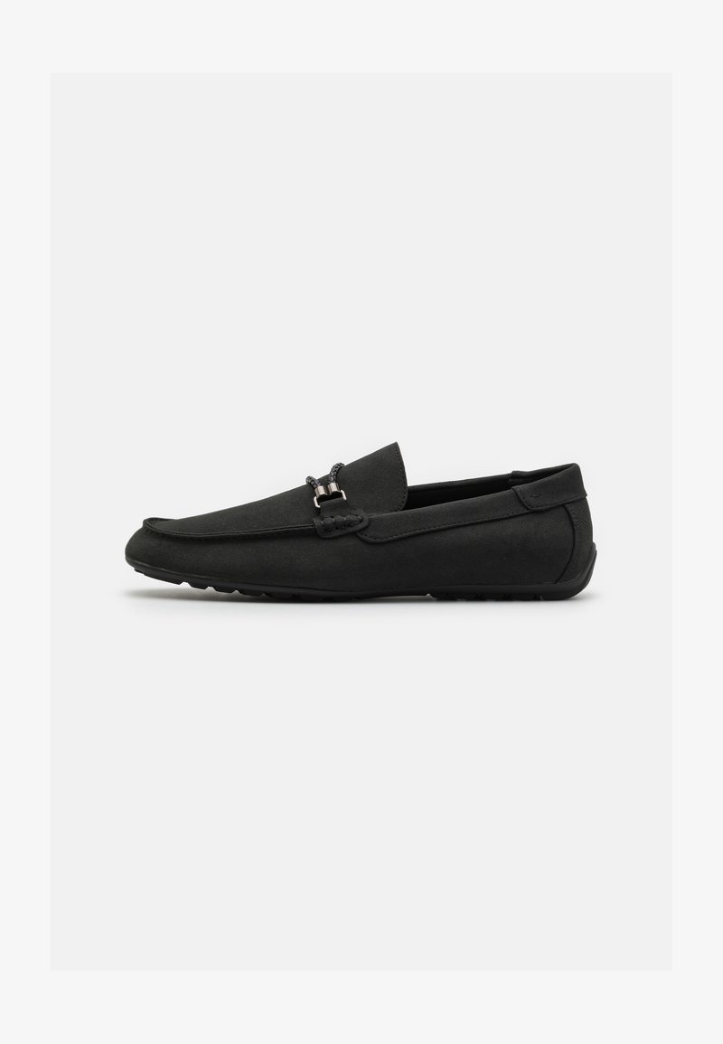 Shoe moccasin preto feito de camurça suave, apresentando uma fivela decorativa na parte frontal, design slip-on e sola de borracha texturizada.
