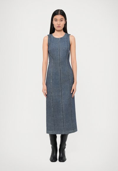 Denim sleeveless dress in dark blue with vertical stitching, fitted design, and a straight silhouette, reaching mid-calf. Black knee-high boots.