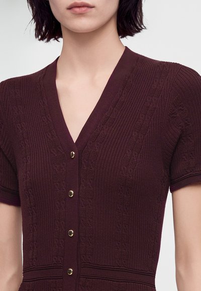 Femme portant un haut en maille violet foncé à manches courtes avec un col en V et quatre boutons dorés sur le devant.