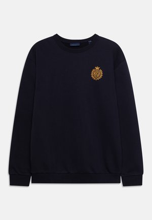 Sweatshirt bleu marine avec col rond, poignets et ourlet côtelés. Présente un blason doré brodé complexe sur la poitrine à gauche.