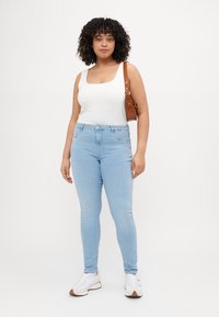 Light blue skinny jeans, white sleeveless top, and white sneakers. Also features a brown crossbody bag with a strap. Plain background.