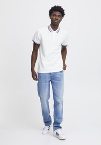 Weißes Poloshirt mit dunkelblauem Kragen und Ärmelausschnitten, kombiniert mit hellblauen Jeans und weißen Sneakers mit schwarzen Streifen.