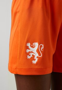 Oranžové športové šortky z textúrovaného materiálu, s bielym logom leva na dolnej strane. Dizajn obsahuje bočný rozparok pre lepší pohyb.