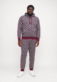 Man in een bijpassende wijnrode en witte geometrische patroon hoodie en joggingbroek met witte sneakers, staand tegen een effen witte achtergrond.