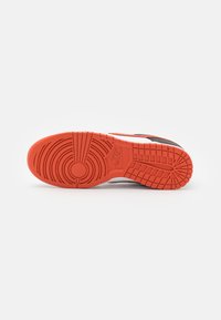 Baskets Nike avec une semelle en caoutchouc présentant un motif texturé orange et des détails circulaires. Des accents noirs et des éléments blancs sont visibles.