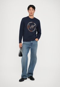 Sweatshirt azul-marinho com um logótipo redondo "MK" em laranja, combinado com jeans claros de perna larga. A pessoa segura uma bolsa preta, posicionando-se contra um fundo branco.