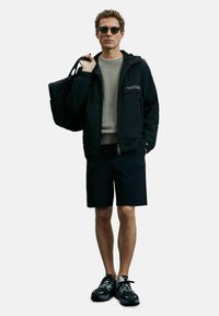 Zwarte waterdichte jas met een kap, grijze gebreide trui, zwarte shorts en zwarte, multifunctionele hardloopschoenen. Inclusief een zonnebril en een zwarte tas.