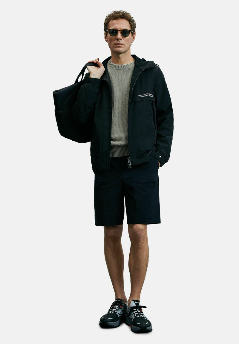 Zwarte waterdichte jas met een kap, grijze gebreide trui, zwarte shorts en zwarte, multifunctionele hardloopschoenen. Inclusief een zonnebril en een zwarte tas.