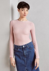 Langärmliges, hellrosa Oberteil mit strukturiertem Muster, kombiniert mit einem hochgeschnittenen, dunklen Jeansrock mit Vorderknöpfen und Taschen.