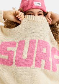 Chunky gebreide trui in beige met grote roze tekst "SURF." Geribde halslijn, zachte textuur en een relaxte pasvorm. Vergezeld van een roze muts.