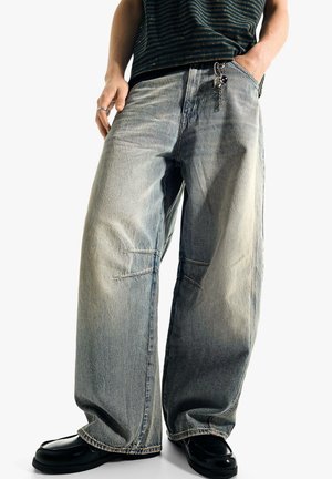 Personne portant un jean bleu délavé et ample avec un porte-clés, des chaussures noires et un haut rayé foncé, debout les mains dans les poches.