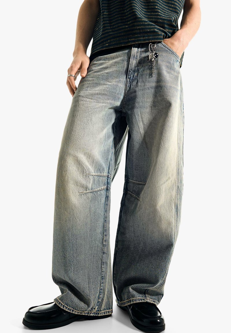 Personne portant un jean bleu délavé et ample avec un porte-clés, des chaussures noires et un haut rayé foncé, debout les mains dans les poches.