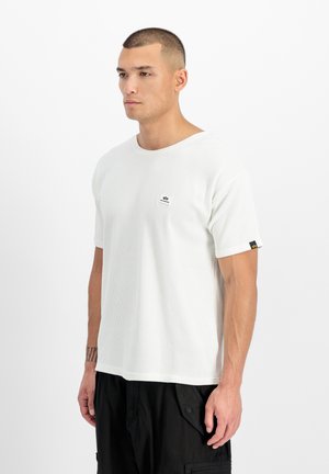 Uomo con capelli corti che indossa una maglietta a maniche corte bianca con trama e pantaloni neri, in piedi davanti a uno sfondo bianco uniforme.