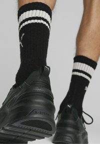 Des chaussures de sport noires avec des semelles texturées, accompagnées de chaussettes noires avec des rayures blanches et un logo en haut.