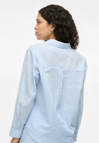 Light blue checkered shirt with long sleeves and button cuffs. Features a classic collar and a back yoke with a vertical seam detail.