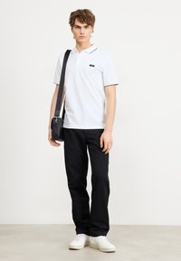 Maglietta polo bianca con bordi neri, abbinata a pantaloni neri e sneakers bianche. Il modello porta anche una piccola borsa a tracolla nera.