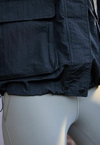 Nahaufnahme einer Person, die eine schwarze, strukturierte Jacke mit großen Taschen und hellgraue, eng anliegende Leggings mit sichtbaren Nähten trägt.