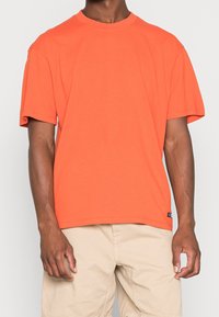 T-shirt a maniche corte in cotone arancione brillante, design a girocollo, vestibilità rilassata, con un piccolo etichetta del marchio sull'orlo inferiore.