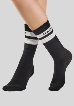 Beine in schwarzen gerippten Crew-Socken mit weißen und schwarzen Streifen und teilweise weißem Text nahe der Spitze vor einem grauen Hintergrund.