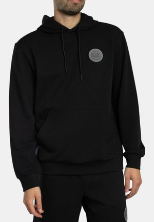 Uomo che indossa una felpa nera con cappuccio e pantaloni abbinati con patch del logo EA7 sul petto e sulla coscia sinistra, in piedi davanti a uno sfondo uniforme.