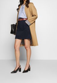 Beige trenchcoat över en ljusblå skjorta med rutiga ärmar, marinblå kjol, brun bälte och svarta slingbackklackar med strukturerad yta.