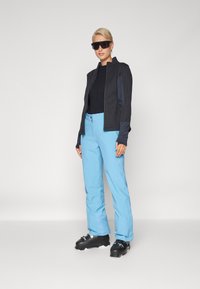 Costum de schi care include o jachetă din material soft-shell de culoare gri închis, un top negru, pantaloni de schi de culoare albastră deschisă și bocanci de schi negri cu catarame.