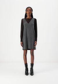 Vestido cinza sem mangas com decote em V, tecido texturizado, sobreposto a uma blusa preta de mangas longas transparente, combinado com botas pretas até ao tornozelo.