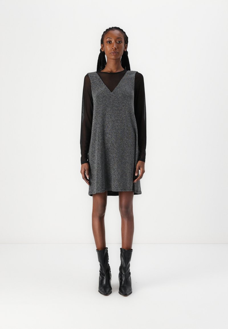 Vestido cinza sem mangas com decote em V, tecido texturizado, sobreposto a uma blusa preta de mangas longas transparente, combinado com botas pretas até ao tornozelo.