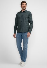 Heren overhemd met lange mouwen in een donkergroene patroonstof, knoopsluiting aan de voorkant, puntige kraag, gecombineerd met lichtblauwe jeans en witte sneakers.