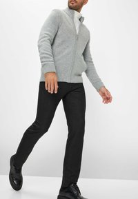 Cardigan zippé gris en matière tricot douce, avec des poignets côtelés et un col montant, associé à un pantalon noir slim et des chaussures noires.