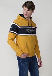 Giovane uomo che indossa una felpa con cappuccio gialla e blu navy con tasca frontale, in posa contro uno sfondo neutro.