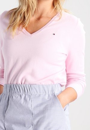 Femme portant un pull rose clair à col en V et un pantalon à taille haute rayé bleu et blanc, les mains dans les poches.