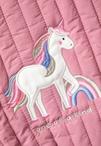 Unicorno bianco con criniera e coda rosa, blu e beige, in piedi su uno sfondo trapuntato rosa con testo ricamato "unico nel suo genere".