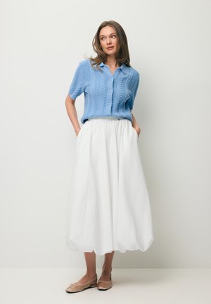 Kobieta ubrana w jasnoniebieski krótki sweter na guziki z krótkim rękawem, białą spódnicę midi z wysokim stanem oraz beżowe baleriny, stojąca z rękami w kieszeniach.