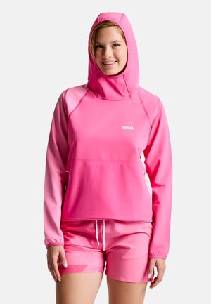 Jeune femme souriante portant un sweat à capuche rose vif et un short rose assorti sur un fond uni.