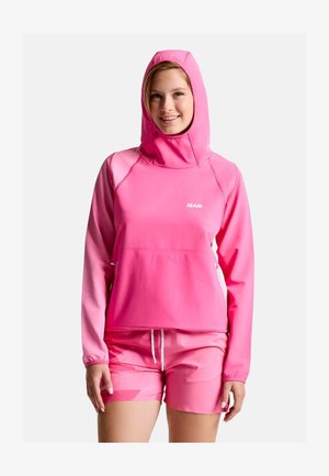 Jeune femme souriante portant un sweat à capuche rose vif et un short rose assorti sur un fond uni.