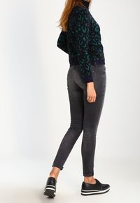 Maglione lavorato a maglia leopardato blu scuro e verde, jeans neri attillati e sneaker neri con suola bianca, mostrato di lato.