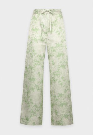Un mannequin se tient devant un fond blanc, vêtu d’un chemisier vert pâle à motifs avec un lien à la taille, d’un pantalon assorti à jambes larges et de chaussures à talons pointus.