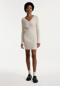 Beiges, eng anliegendes Rippkleid mit langen Ärmeln, V-Ausschnitt und asymmetrischem Wickeldesign. Kombiniert mit schwarzen Plateauschuhen und weißen Socken.